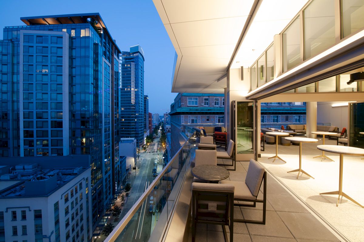 A modern rooftop terrace with seating overlooks a cityscape at dusk, featuring tall buildings and illuminated windows.