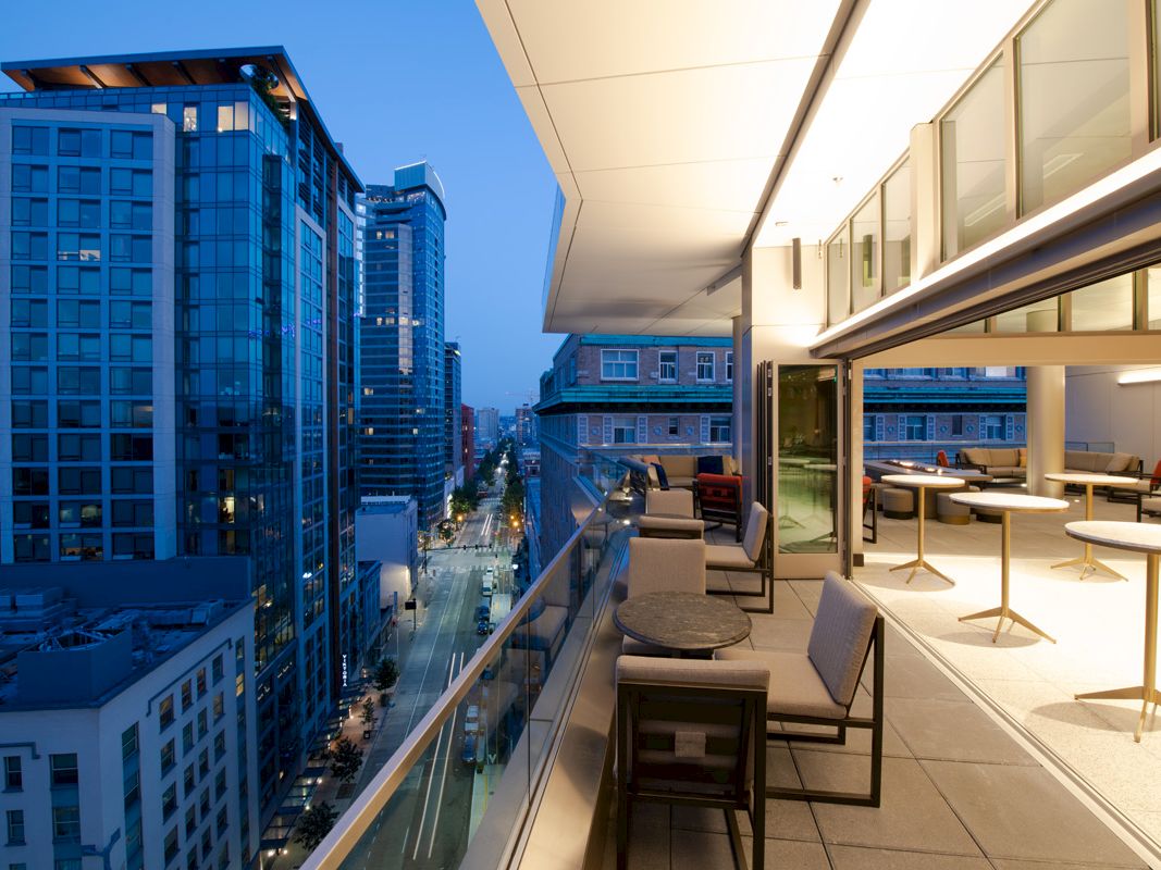 A modern rooftop terrace with seating overlooks a cityscape at dusk, featuring tall buildings and illuminated windows.