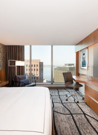 A modern hotel room features a large bed, flat-screen TV, work desk, and a window with a city view, showcasing a sleek and cozy design.