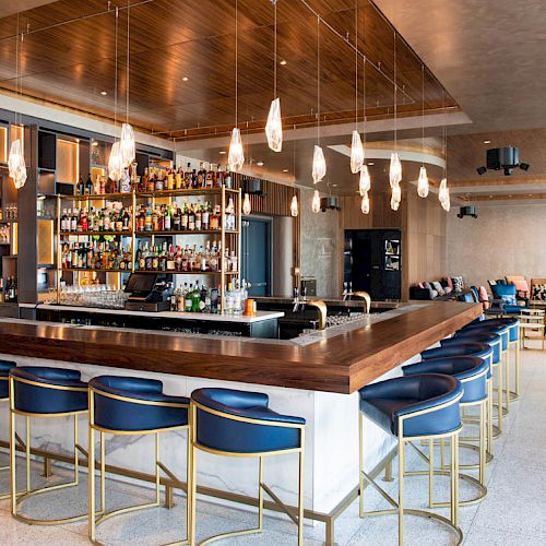 A modern bar with blue stools, hanging lights, and a fully stocked display surrounded by sleek, contemporary decor and seating areas.