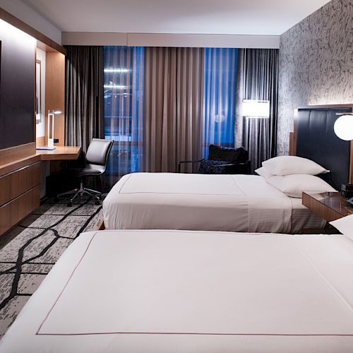 The image shows a modern hotel room with two beds, a TV, a work desk, and lamps, featuring stylish decor and cozy lighting.