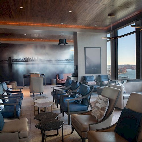 This image shows a modern lounge with seating, tables, large windows, and a coastal view. A wall mural features a cityscape.