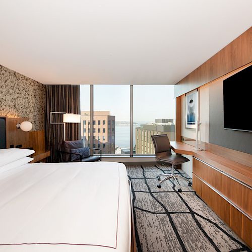 A modern hotel room features a large bed, a flat-screen TV, a desk with a chair, and a window with an urban view.