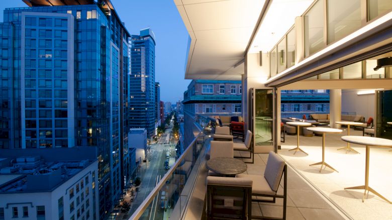 A modern cityscape with high-rise buildings at dusk, featuring a stylish rooftop patio with seating and tables along the terrace.