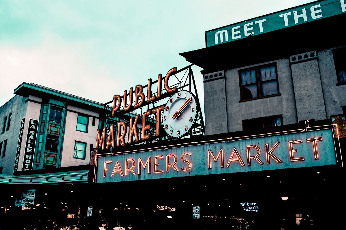 Neon signs for "Public Market" and "Farmers Market," with an iconic clock, are visible on buildings under a cloudy sky.