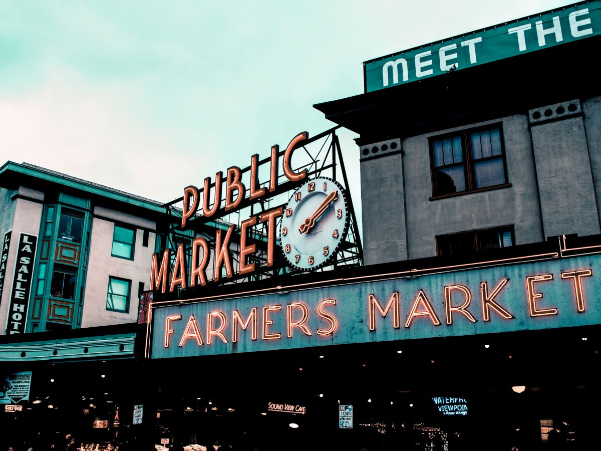 Neon signs for "Public Market" and "Farmers Market," with an iconic clock, are visible on buildings under a cloudy sky.