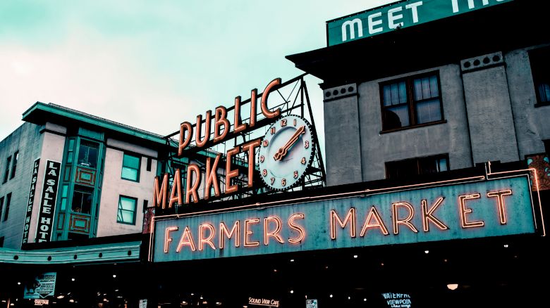 Neon signs for "Public Market" and "Farmers Market," with an iconic clock, are visible on buildings under a cloudy sky.