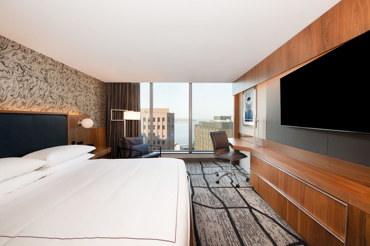 A modern hotel room with a large bed, wall-mounted TV, desk, chair, and window with a cityscape view.