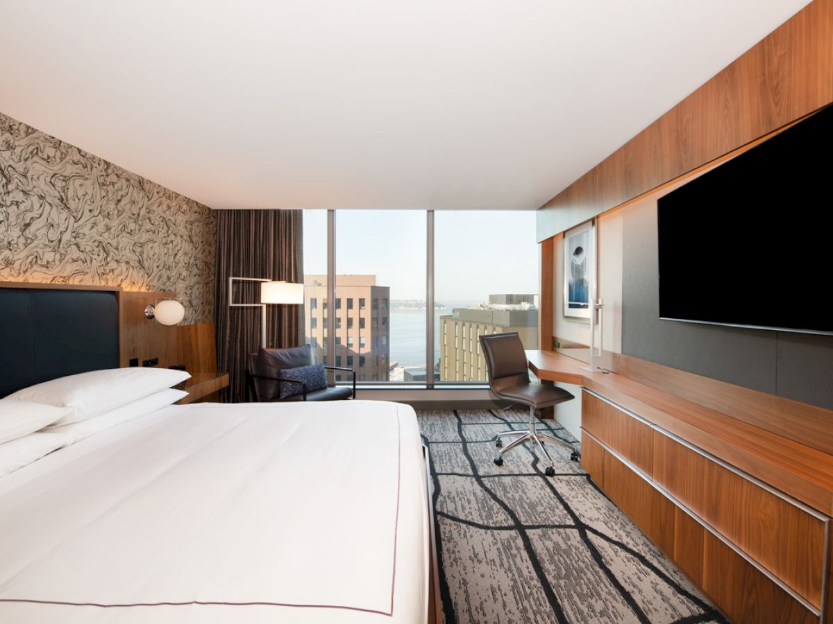 A modern hotel room with a large bed, wall-mounted TV, desk, chair, and window with a cityscape view.