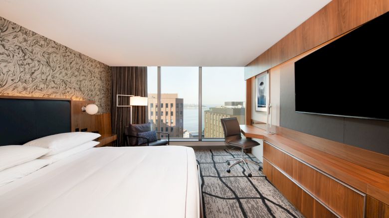 A modern hotel room with a large bed, wall-mounted TV, desk, chair, and window with a cityscape view.