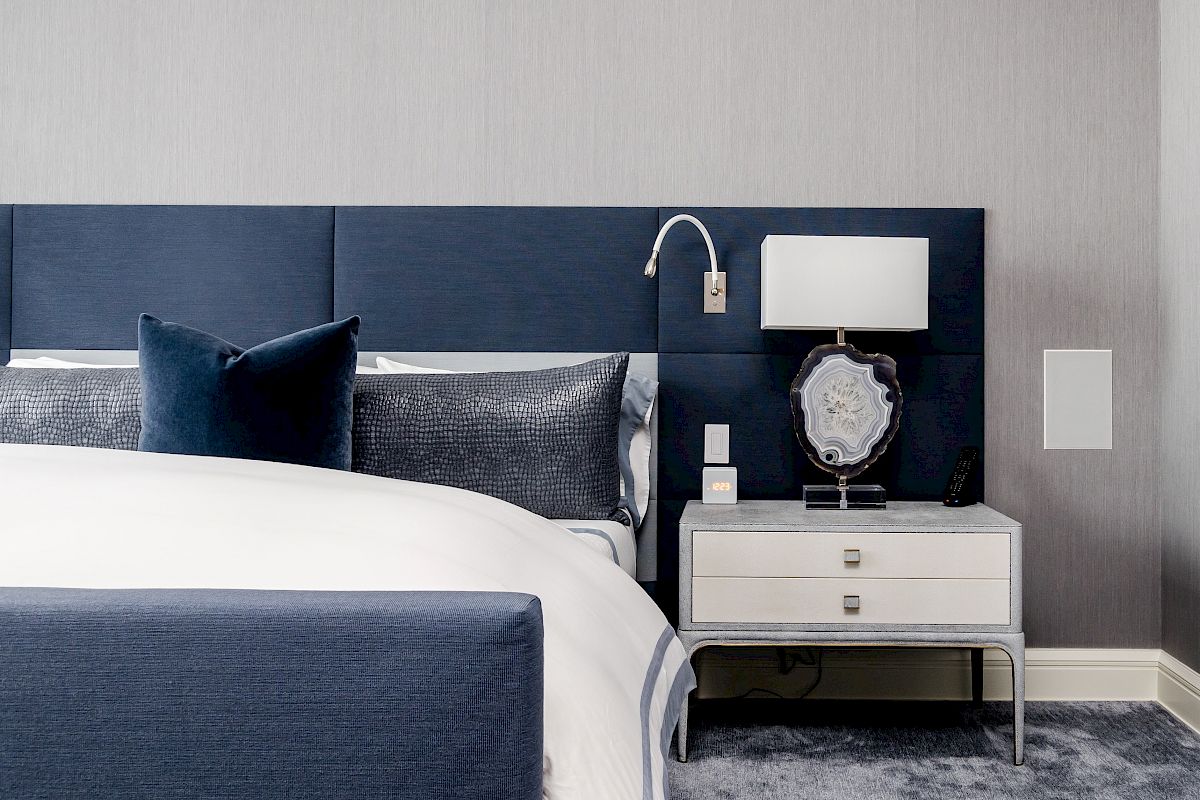 A modern bedroom with a blue headboard, white nightstand, lamp, and framed art piece. Clean and minimalist design.