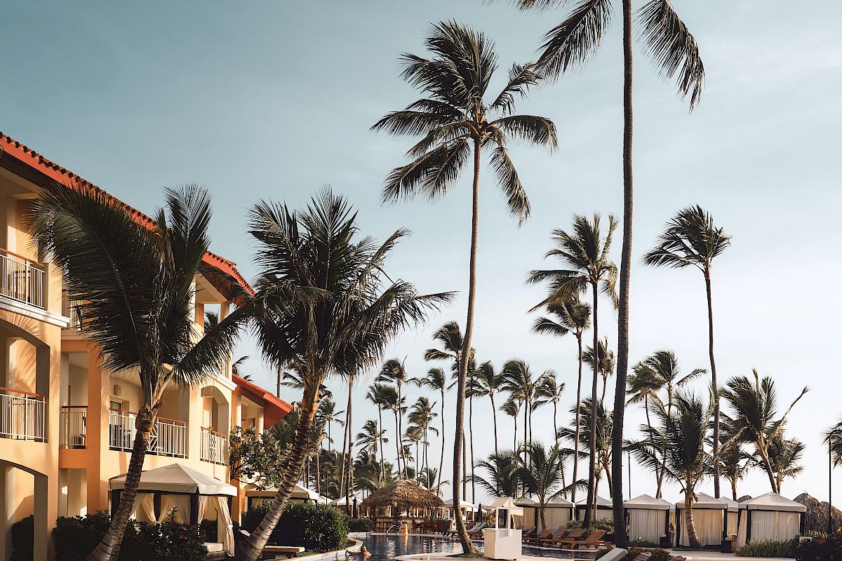 A tropical resort scene with a pool, lounge chairs, palm trees, and buildings under a partly cloudy sky at sunset.