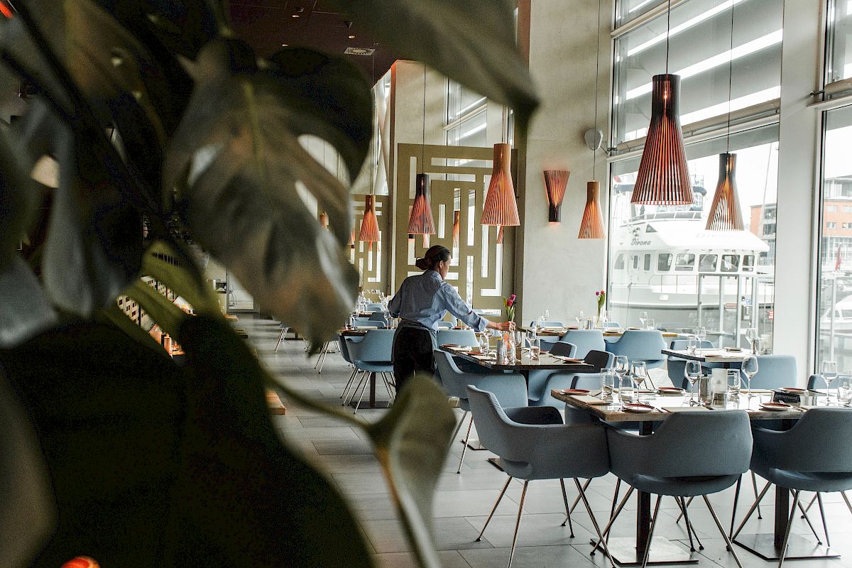 A modern restaurant with elegant chandeliers and a person setting tables, visible through large windows and plant decor in the foreground.