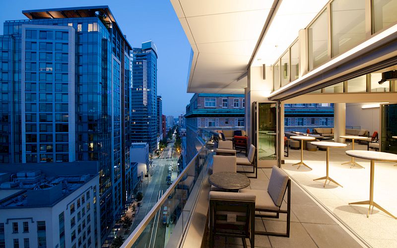 A modern rooftop terrace at dusk, overlooking city buildings. Cozy seating and tables are arranged for relaxation or gathering.
