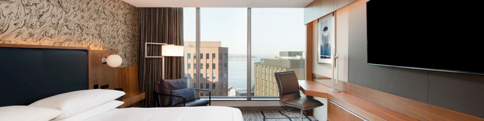 A modern hotel room with a large bed, flat-screen TV, desk, chair, and window view of city buildings and waterfront.