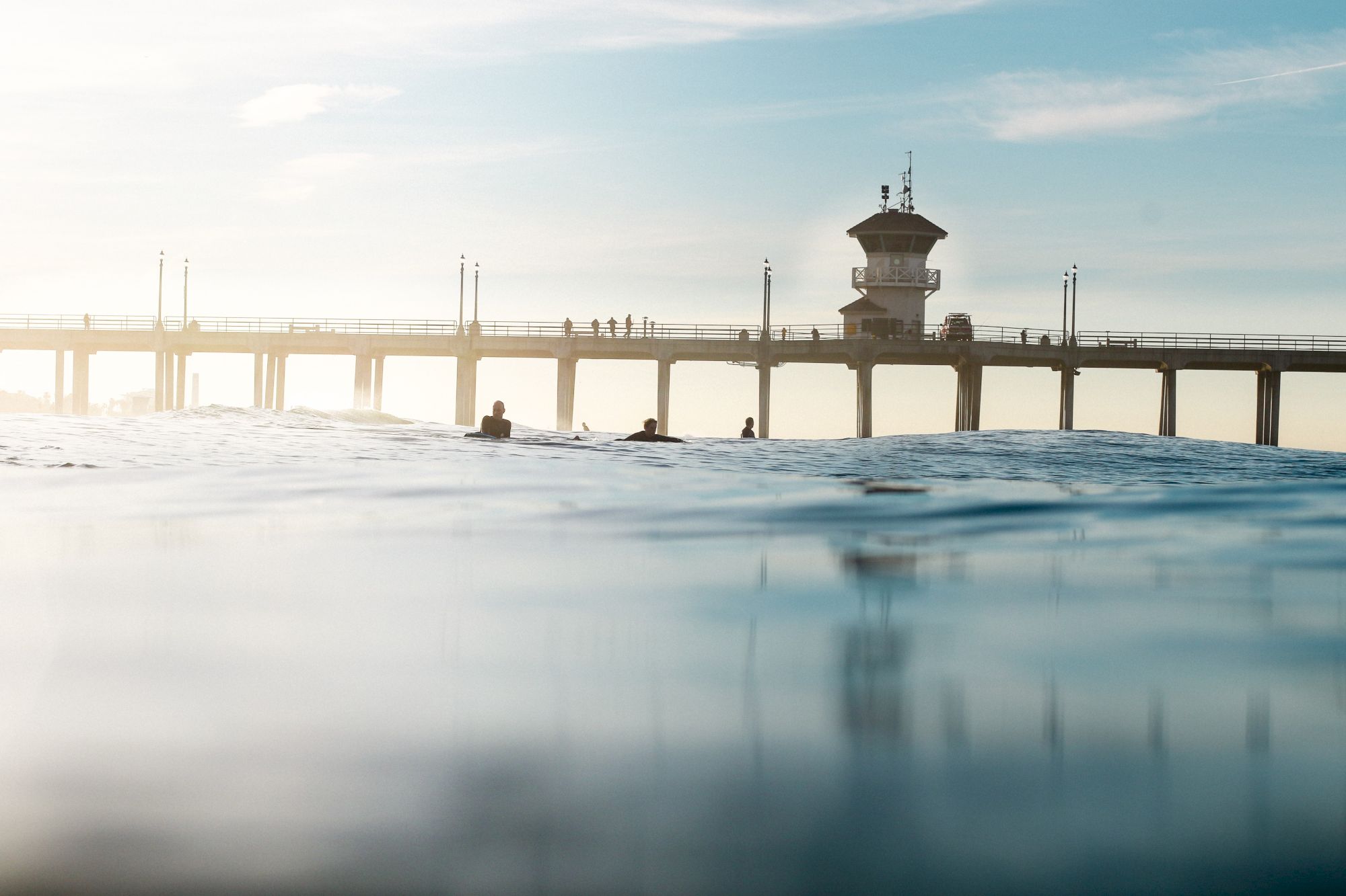 The image shows a pier extending over calm water with people walking on it under a clear sky.